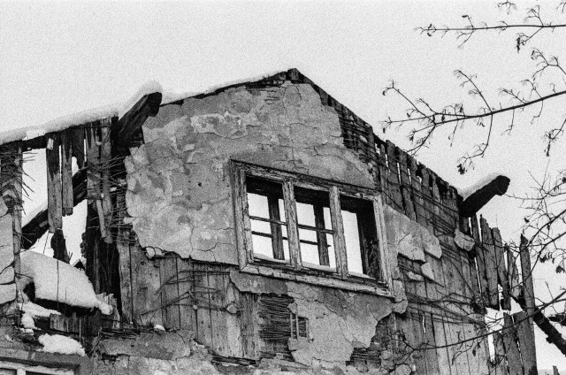 The image shows a dilapidated, weathered building in black and white. The structure appears to be in a state of significant disrepair, with large portions of the walls crumbling and the wooden framework exposed. A broken window with old, peeling frames is visible, and snow is accumulating on parts of the decayed wooden beams. The scene has a bleak, almost abandoned atmosphere, heightened by the stark contrast and grainy texture of the image. Bare tree branches reach into the frame from the right side, adding to the overall desolate mood.