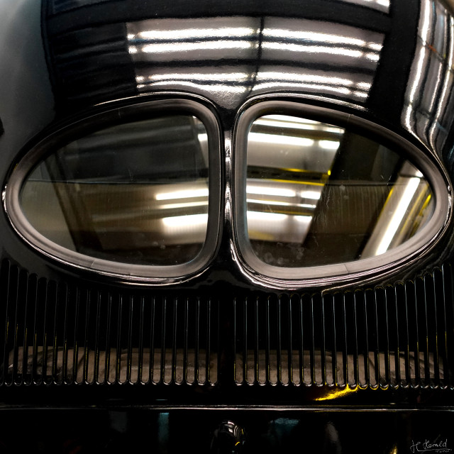 Close-up of a split rear window of an old black VW Beetle. Ceiling lights and some yellow accents are reflected in the windows and roof.