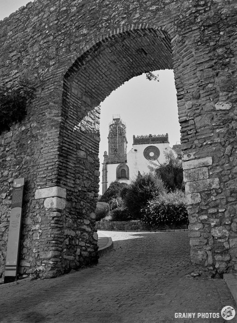 A black-and-white film photo of the Santa María la Mayor church, viewed through the arch of the town entrance in the town walls.