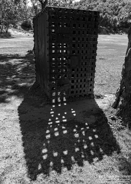 A black and white portrait photo of an old outdoor iron jail "hot box." Wide iron rails form a tight lattice around all four sides and the top. It's small, probably meant for one person. It casts a wide pyramid shaped shadow in the foreground. A tree trunk is seen on the right. A grassy field is in the background with heavy trees and brush in the distance. 