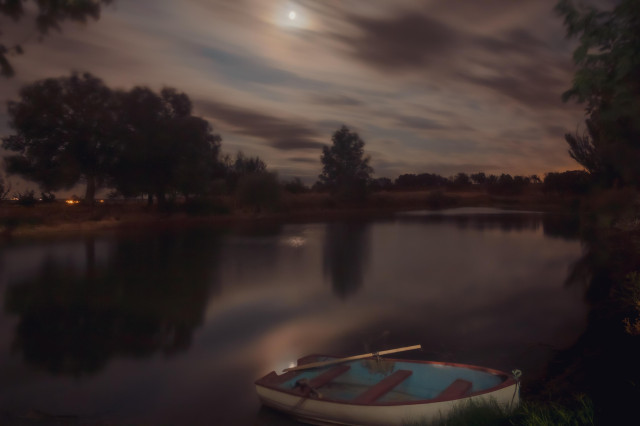 Fotografía nocturna donde se ve una balsa con una barca en primer término y un cielo nocturno con nubes y luna llena entre ellas.