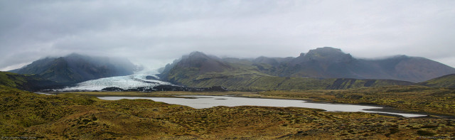 A wide photo of a mountain range under low cloud. The foreground is brown vegetation across lumpy ground. Just beyond that is a pool of greyish water - meltwater from glacial activity usually carries fine particles of rock. Across the centre of the shot are three distinct peaks, their heights shrouded in thick cloud. Between the leftmost and the next peak to its right, a tongue of ice is dropping to ground level, the origin of which is hidden in the mists.