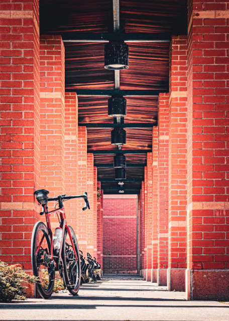 Photo of a red BMC roadmachine road bike, leaning up against column made of red bricks in an outdoor setting. The column is one of many that are in two rows, and we are looking straight down the middle of both those rows. There is a roof joining the two columns that is made of narrow wooden planks. The roof has a row of hanging lights running right down the middle of it. 