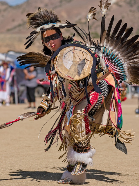 Color portrait photo of a Native American powwow dancer. He wears buckskins that are decorated with numerous bones and feathers. He carriers a leather shield on the face of which is the image of a Native with long black hair shooting a bow and arrow into the air. The dancer's face is painted with a black stripe from ear to ear across his eyes and two thin white stripes vertically on the right side of his face. 