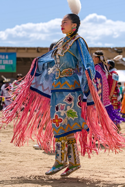 Color portrait photo of a female Native American powwow dancer. She wears light blue regalia with a shawl that has long dark pink fringe. The regalia is beaded with images of leafs, flowers, a gold zigzag design around her waist, an elaborately beaded belt of flowers and leafs. Her tall moccasins are beaded in white, yellow, blue, and red geometric and botanical designs. She wears a fluffy white plume at the back of her head. Her hair is black and braided. She is expressionless and in profile to the camera, standing straight, but both feet are off the ground. 