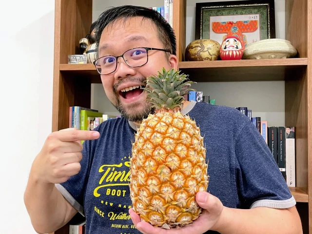 A man happily holding a ripe yellow pineapple in his left hand, while pointing at the pineapple with his right hand, smiling at the camera.