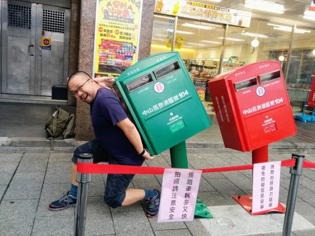 A man kneels down next to two tilted mailboxes in Taipei, Taiwan, pretending to be carrying one of the mailboxes on his back.