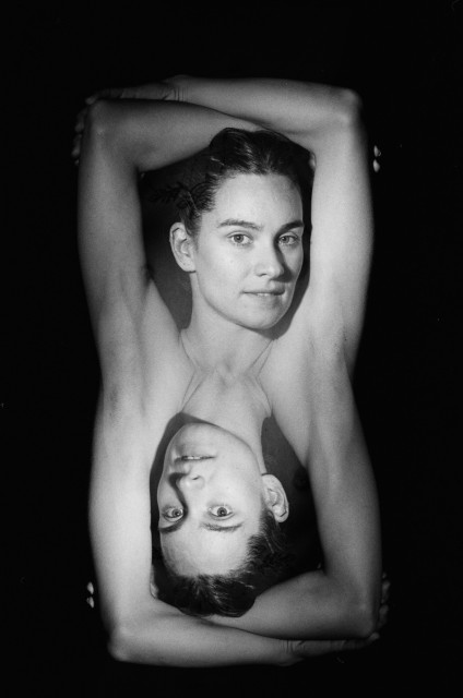 A woman's shoulders and arms are doubled through the double exposure so that you see two of the same woman upside down, joined at the armpits. Her arms are above her head in both images. Black and white. 