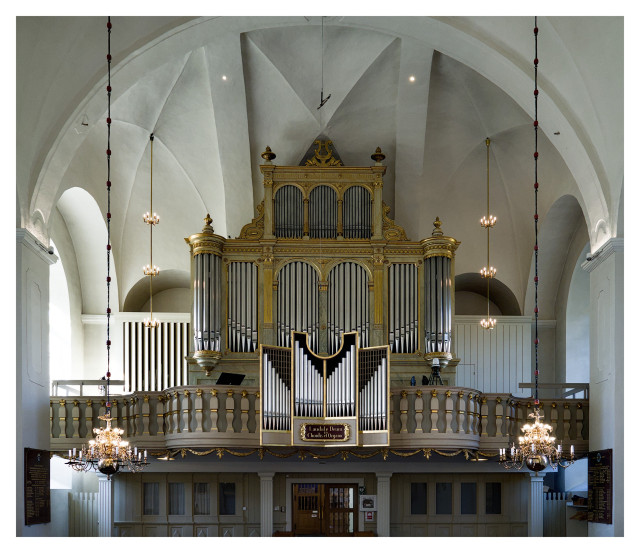 En kyrkoorgel ovanför en dörr och en rad fönster. Piporna är silverfärgade med guldfärgade detaljer mellan delar. Kyrkans vita stenvägg som bakgrund.