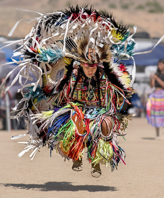 Color portrait photo of a Native American "Fancy" dancer at a powwow. Fancy dancers have copious amounts of colorful ribbons tied to their regalia. The prominent colors in this photo are: white, green, red, orange, yellow, and a little blue. This dancer has jumped and both feet are tucked up and off the ground. She has long black braided ponytails and a black shawl across her shoulders and upper arms. She is slightly bent forward, in the tucked jump, and looks down toward the ground.