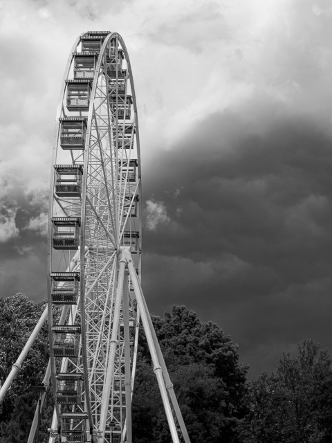 Hochformat. Ein Riesenrad, Schmalseitenansicht. Dahinter eine dunkle Wolke.