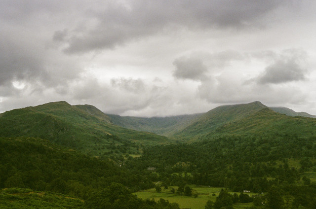 A lush green mountain range in the shape of a horseshoe, with woods and houses in front of it, underneath a cloudy grey sky.