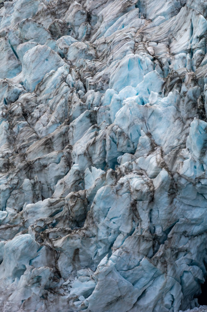 A detailed view of a chaotic jumble of ice seracs at the Gabriel Icefall on the Gulkana Glacier in the Eastern Alaska Range. The image shows a mass of jagged, irregularly shaped ice formations with varying shades of blue, interspersed with dark streaks of sediment and debris, completely filling the frame. The seracs appear densely packed, with intricate layering and fractures.