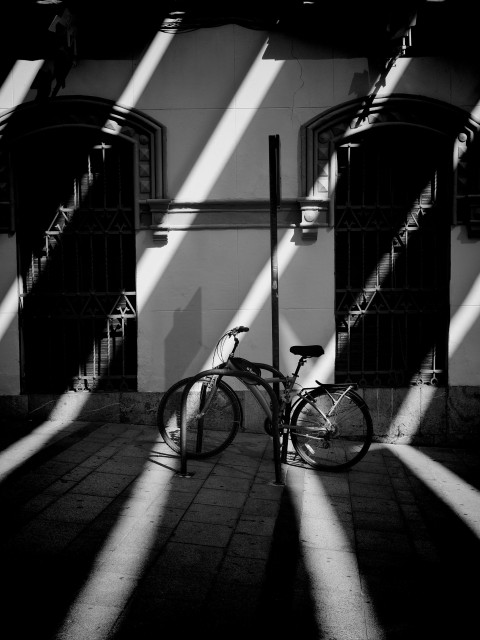 A bicycle leaning against a white building in Córdoba's historic district where overhead shades are casting diagonal stripes of bright sunlight and dark shadows across the scene