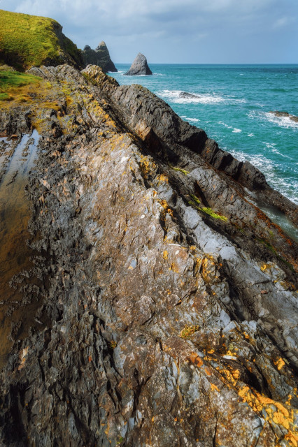 This striking image showcases the jagged, rocky shoreline of the Pembrokeshire coast. In the foreground, weathered rock formations dominate, their surfaces mottled with patches of grey, gold, and rusty orange lichen. The rocky outcrops stretch diagonally across the frame, leading the eye towards the turquoise sea beyond. In the distance, a dramatic sea stack rises from the water, its pointed silhouette stark against the horizon. The grassy cliff tops are visible to the left, providing a soft contrast to the harsh, angular rocks. White-capped waves crash against the shore, emphasising the rugged beauty and raw power of this windswept coastal landscape.
