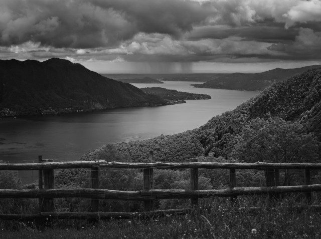 Schwarz-weiß Foto eines Sees in einer Berglandschaft. Der See führt vom linken Bildrand in die Bildmitte und schlängelt sich durch die Berglandschaft.
Im Vordergrund befindet sich ein Holzzaun in einer Wiese.
Der Himmel ist bewölkt und in der Ferne, am Seeende ist ein Regenschauer erkennbar.