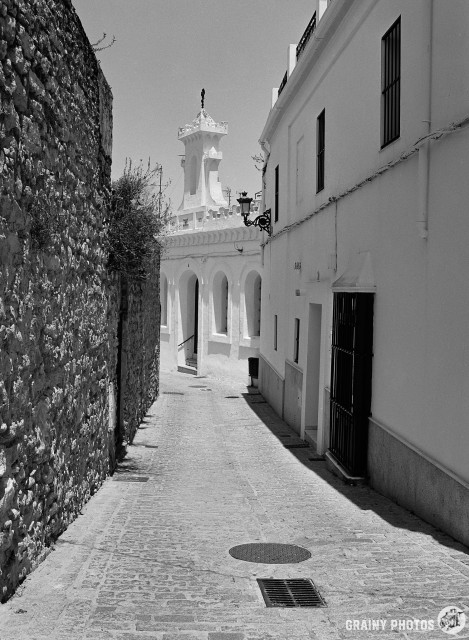 A black-and-white film photo of a narrow cobbled street. On the right is a white house and a small chapel with a belfry. A stone wall is on the left