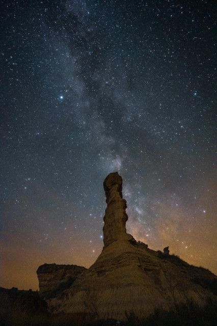 Se puede ver una formación rocosa desértica con forma alargada hacía un cielo estrellado con la Vía Láctea sobre ella.