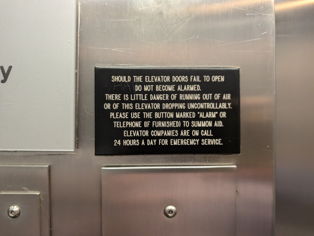 A black, engraved plastic sign in an elevator. It reads:

SHOULD THE ELEVATOR DOORS FAIL TO OPEN
DO NOT BECOME ALARMED.
THERE IS LITTLE DANGER OF RUNNING OUT OF AIR
OR OF THIS ELEVATOR DROPPING UNCONTROLLABLY.
PLEASE USE THE BUTTON MARKED "ALARM" OR
TELEPHONE (IF FURNISHED) TO SUMMON AID.
ELEVATOR COMPANIES ARE ON CALL
24 HOURS A DAY FOR EMERGENCY SERVICE.