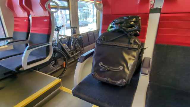 Blick zwischen zwei Bahnsitzen in rot und anthrazit durch.
Man sieht ein graues Fahrrad hinten und eine schwarze Fahrradtasche am Sitz im Vordergrund