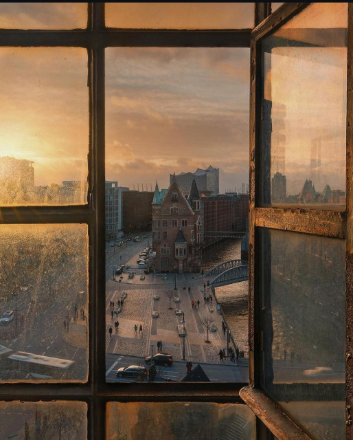 Photography. A color photo with a view of the city of Hamburg from a window. The dark foreground shows the view through a historic, old wooden window consisting of 12 small panes. The middle part of the window, which is divided into two panes, is open and provides a view of the city in orange sunlight. The sun falls over the city from the left and bathes the streets in a yellow-orange light, which is also reflected in the window panes. You can see streets with a few illuminated cars and a few passers-by walking around. The direct view shows the tall old brick buildings of the "Speicher-Stadt", an elongated, historic warehouse complex in the port of Hamburg with canals and bridges. In the distance, as if in contrast, you can see the modern glass façade of the Elbphilharmonie concert hall.  Above it is a slightly cloudy yellow sky. Like a glimpse from the past into the future and the knowledge that this special light has always been there.