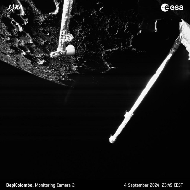 A black-and-white picture of part of the planet Mercury showing lots of craters and other surface features, seen against a black sky with some parts of the ESA/JAXA BepiColombo spacecraft in the foreground.