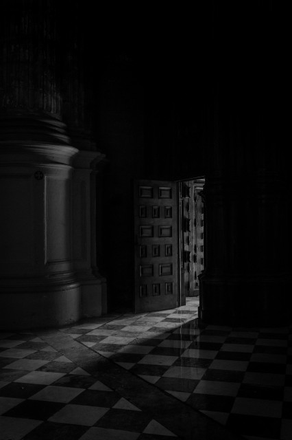 Fotografía en blanco y negro en formato vertical donde se ve una puerta de la catedral de Málaga por la que entra algo de luz e ilumina un poco el interior pero sigue habiendo bastantes sombras.