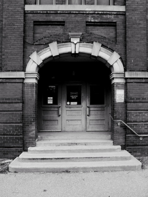 Three large doors under an archway of an old school can be seen in this black and white image.