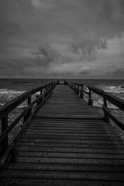 Das Foto zeigt einen alten, defekten Steg der in die Ostsee hineinragt. Der Himmel ist stark bewölkt, das Meer rechts und links des Stegs ist unruhig. Das Foto ist in schwarzweiss. 