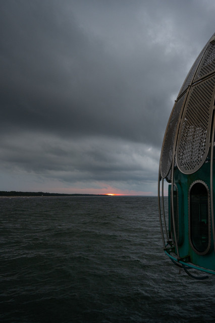 Das Foto zeigt die Tauchglocke auf der Seebrücke in Zingst. Der Himmel ist stark bewölkt, Wellen zeigen sich auf dem Meer. Ganz im Hintergrund ist ein Hauch des Sonnentuntergangs zu sehen. Eher ein kleiner roter Punkt am Horizont.