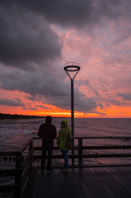 Das Foto zeigt 2 Menschen auf der Seebrücke in Zingst, die unter einer Laterne stehen und auf das Meer hinausschauen. Die Sonne geht unter und zeigt sich in den orangeroten Tönen, aber darüber sind tiefschwarze Wolken zu sehen. Die ganze Szenerie bekommt dadurch ein sehr spezielle Stimmung.