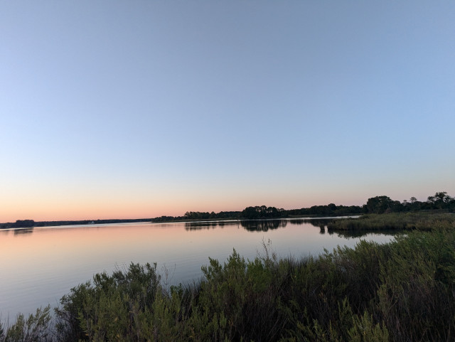 Photo of a calm late summer morning on Harris Creek sun rising just below the horizon and illuminating it bright orange. Sky is deep blue and cloudless. Marsh is green and ready for the day. Creek water is flat calm and perfectly reflects the sun and sky.