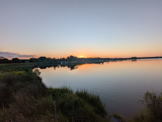 Photo of a calm late summer morning on Harris Creek sun rising just below the horizon and illuminating it bright orange. Sky is deep blue and cloudless. Marsh is green and ready for the day. Creek water is flat calm and perfectly reflects the sun and sky.