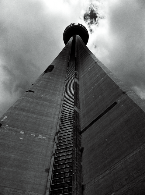 Black and white photo looking up at the very tall concrete CN Tower in Toronto, Ontario, Canada. The background is a drak cloudy sky with a small break in the clouds visible at the top of the frame.