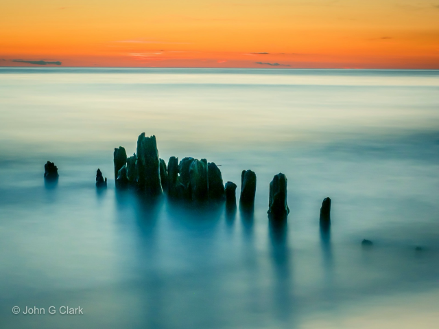 Bits of old pier piling in blue smooth water with an orange sunset in the back