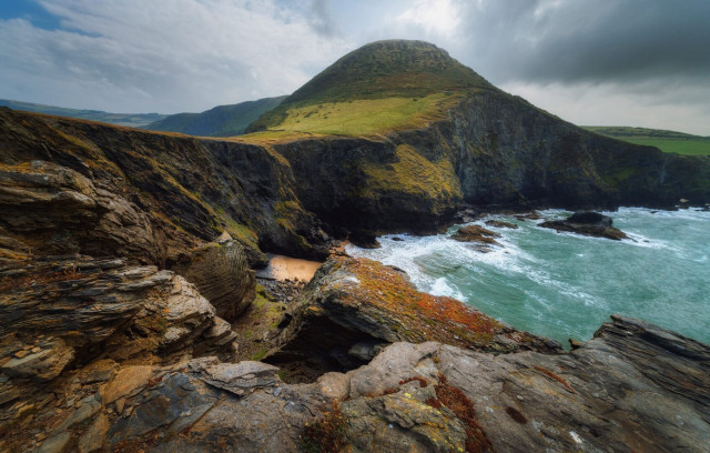 The rugged beauty of the Ynys Lochtyn peninsula near Llangrannog, Wales. In the foreground, jagged rock formations create a dramatic coastline, their layered striations telling tales of ancient geological processes. A small, secluded sandy cove is nestled amongst the cliffs, its golden sands a stark contrast to the dark, craggy rocks surrounding it. The sea crashes against the shore, its turquoise waters frothing white as they meet the land. Dominating the scene is a majestic, verdant hill rising from the sea, its slopes a patchwork of lush greens and earthy browns. The sky above is a canvas of swirling clouds, lending an air of moody grandeur to this breathtaking Welsh landscape.