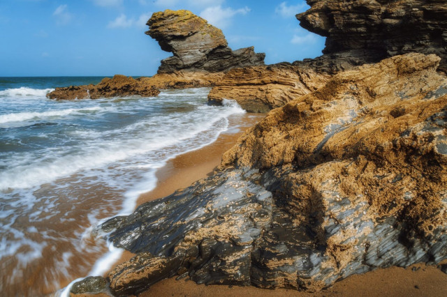 This image captures a dramatic coastal scene at Llangrannog near Cardigan in Wales. Rugged, golden-hued cliffs dominate the foreground, their layered rock formations sculpted by time and the elements. A large, distinctive rock formation known as Carreg Bica juts out from the cliff face, resembling a sentinel overlooking the sea. The shoreline is a mix of sandy beach and rocky outcrops, with foamy waves gently lapping at the shore. The sea stretches out to meet a clear blue sky dotted with wispy clouds, creating a serene backdrop to the raw beauty of the Welsh coastline.