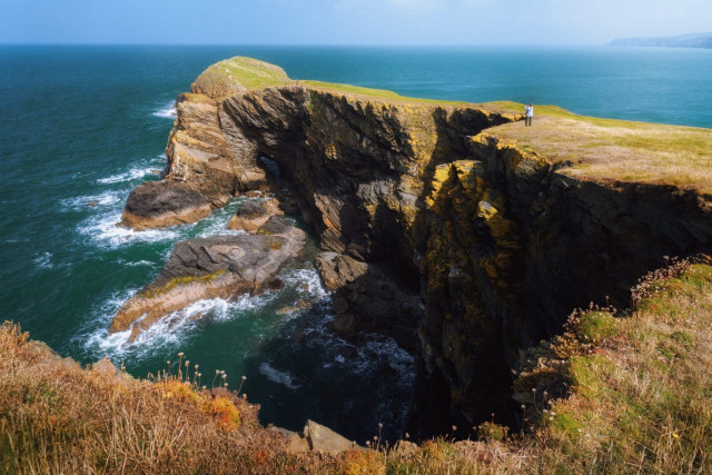 The dramatic coastal landscape of Ynys Lochtyn peninsula near Llangrannog, Wales. Towering cliffs of layered rock face the deep blue-green sea, their rugged faces carved by millennia of wind and waves. The cliff tops are adorned with lush grass and wildflowers, creating a stark contrast to the dark, craggy rock below. White-capped waves crash against the base of the cliffs, highlighting the raw power of the ocean. In the distance, the coastline stretches on, fading into a misty horizon. A lone figure stands near the cliff edge, providing scale and emphasising the majestic grandeur of the scene. The image captures the wild, untamed beauty of the Welsh coast, with its interplay of land, sea, and sky.