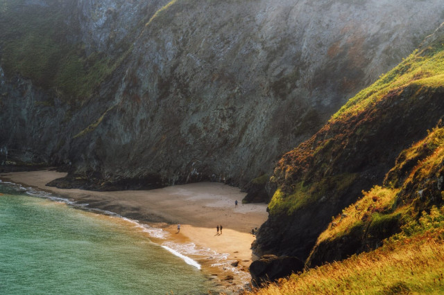 A secluded beach nestled between towering cliffs at Ynys Lochtyn peninsula near Llangrannog, Wales. The golden sandy shore is embraced by imposing grey rock faces, their sheer height emphasised by the tiny figures of beachgoers below. Sunlight bathes the right-hand cliff in a warm glow, illuminating the verdant vegetation that clings to its slopes. The sea, a mesmerising turquoise, laps gently at the shore, creating a serene contrast to the rugged landscape. The scene captures the raw beauty of the Welsh coastline, where dramatic geology meets tranquil waters, offering visitors a sense of awe and isolation amidst nature's grandeur.