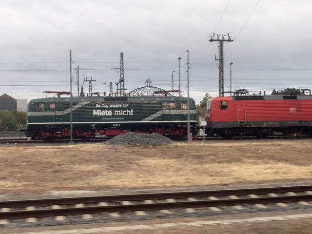 A DB 143 locomotive that’s green with “Miete mich” written on it. The loco behind it is plain and red. 