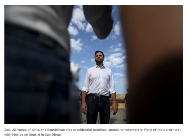 A screenshot from an NPR article. A photograph is embedded showing JD Vance standing in the center of the frame. Two reporters flank him on either side in the foreground, made blurry by the camera focus. One of the reporters arms sticks out in the middle, making the general shape of something... suggestive... pointing at Vance.