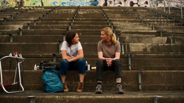 Cailee Spaeny and Kirsten Dunst sitting and talking in a run down stadium.