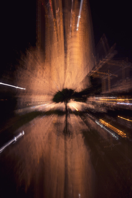 Fotografía nocturna y vertical donde se deforma mediante el movimiento de zoom de larga exposición la imagen de la Sagrada Familia y su reflejo.