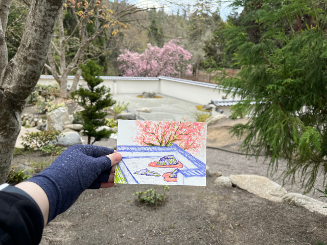 Japanese garden in the spring! A hand in a compression glove holds out an artwork in front of its inspiration, a walled garden with rocks, and a blossoming tree visible behind it, in bright, somewhat fantasy-like, tertiary colors.