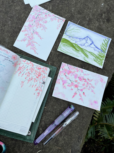 Three artworks and a planner, one artwork of Mount Hood in lavender and green, two of cherry blossoms in peachy pink. The pens used for the cherry blossoms lie beside the postcard size original artworks. The right two (Mt Hood, cherry blossom diagonal) are still available for sale!