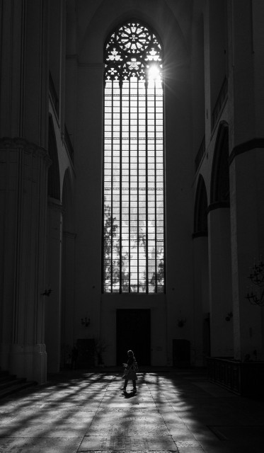 Das Fenster zeigt eine riesiges Fenster in der 
Marienkirche in Stralsund. Der obere Teil ist mit Mustern verziert. 
Die Rahmen des Glases werfen Schatten auf den Boden. Unten ist noch eine Frau zu erkennen, sie wirkt sehr klein.
Oben rechts zeigt sich noch die Sonne in dem Fenster. Das Bild ist in schwarzweiß. 