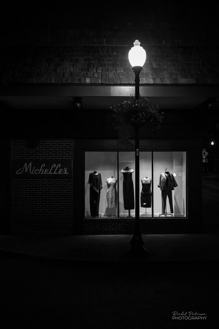 A lighted street display window for a women's clothing store is shown at night where the only lighting is the display lights and one streetlight.