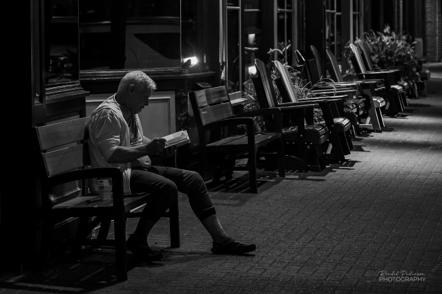 A man sits at night on a bench reading a book and smoking a cigarette on the sidewalk of historic Saint Charles, Missouri.