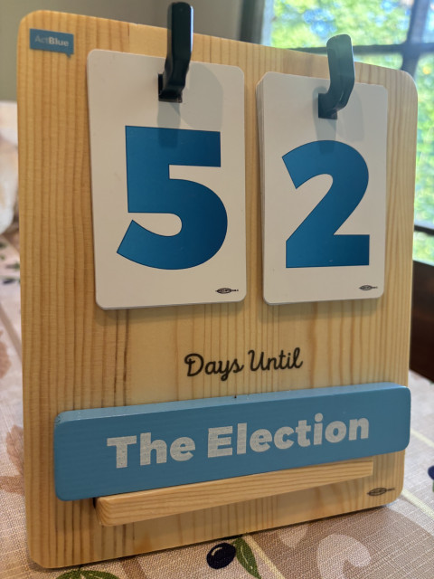 A wooden countdown board displays "52 Days Until The Election" with numbers and text in blue on a natural wood background.