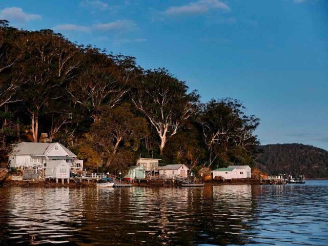 A serene waterfront scene with several houses nestled among trees, reflecting in the calm water under a blue sky.